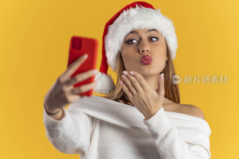美丽的年轻女子戴着圣诞老人的帽子，对着她的智能手机相机飞一个吻，在黄色背景上进行视频通话/自拍